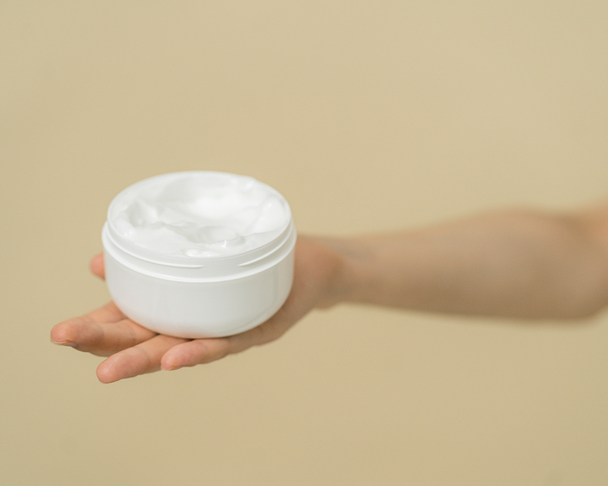 Domáci pleťový krém v bielej plastovej nádobe drží ženská ruka pred béžovým pozadím.